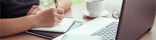 Image of someone writing on a notebook, with an open laptop in front of them and a coffee cup on the side.